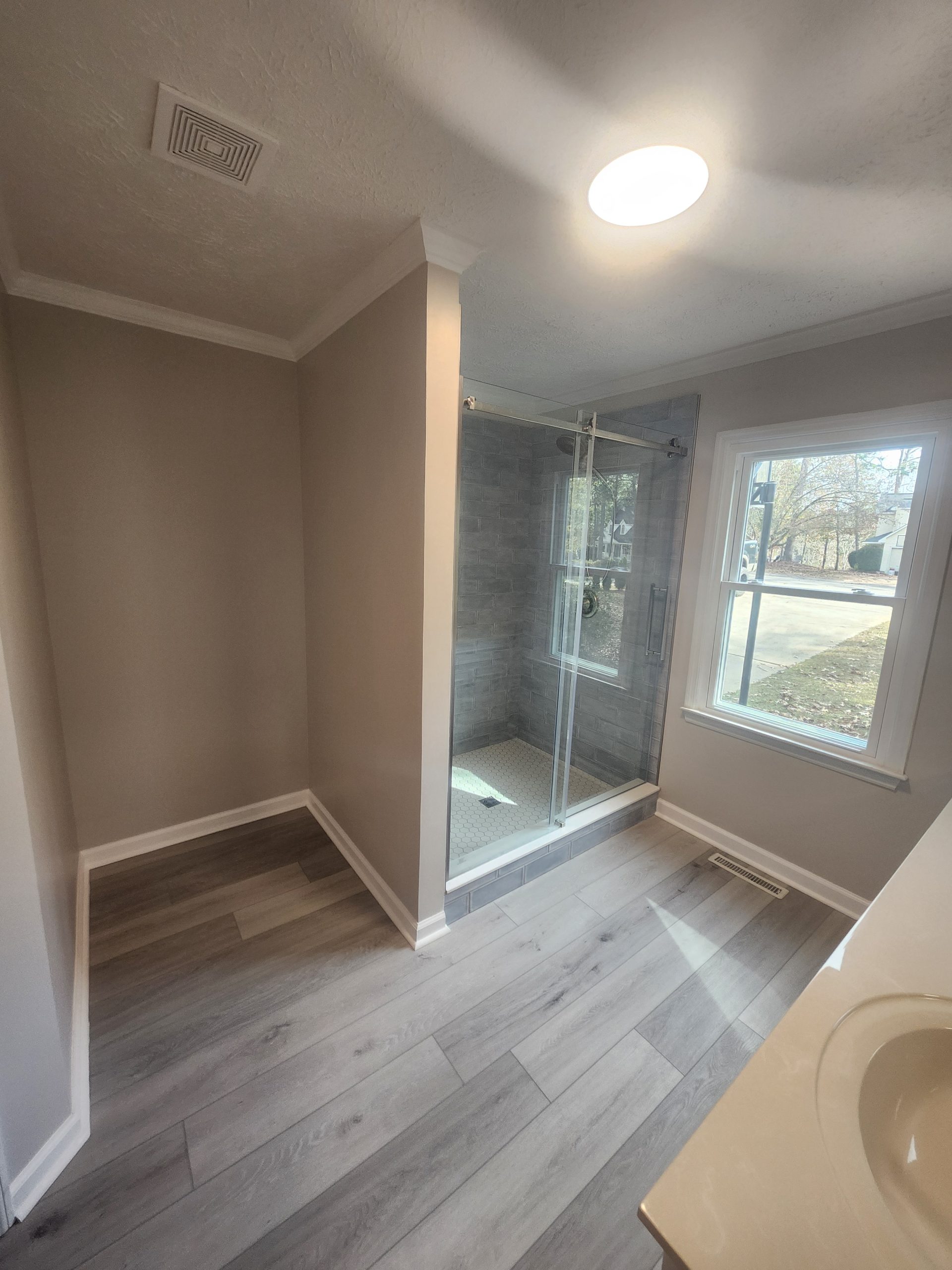 Modern master bathroom renovation featuring walk-in shower with gray tile and glass enclosure, light wood-look plank flooring, and recessed lighting.