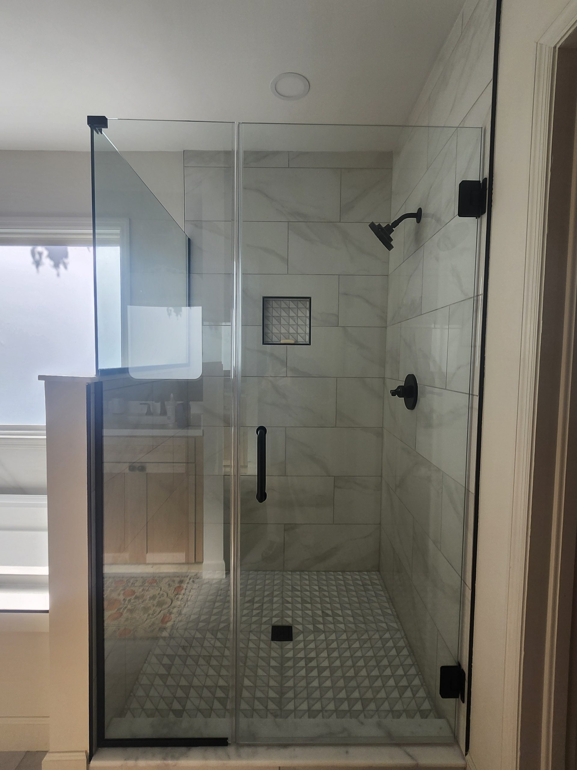 Frameless glass walk-in shower with light gray subway tile walls, textured floor tile, and black fixtures installed by bathroom renovation professionals.