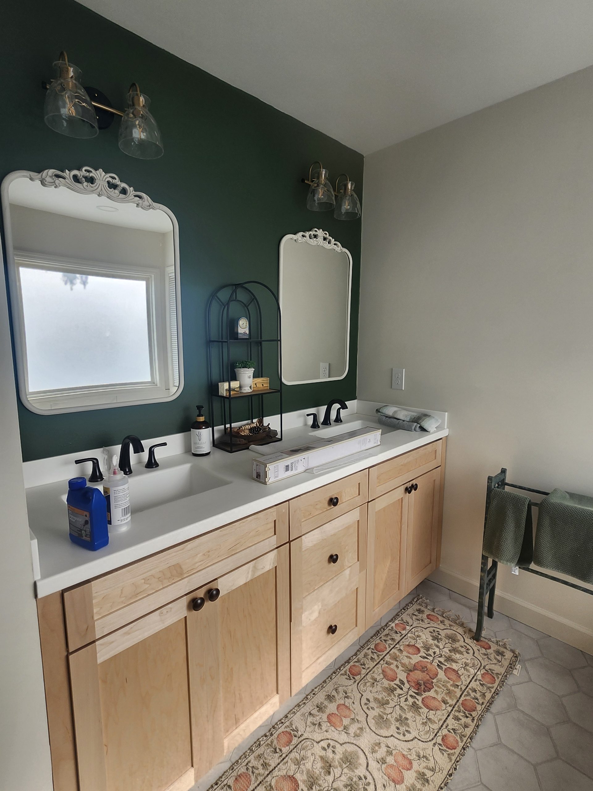Dual-sink bathroom vanity with light wood cabinetry, white countertop, forest green accent wall, and brass pendant lighting.