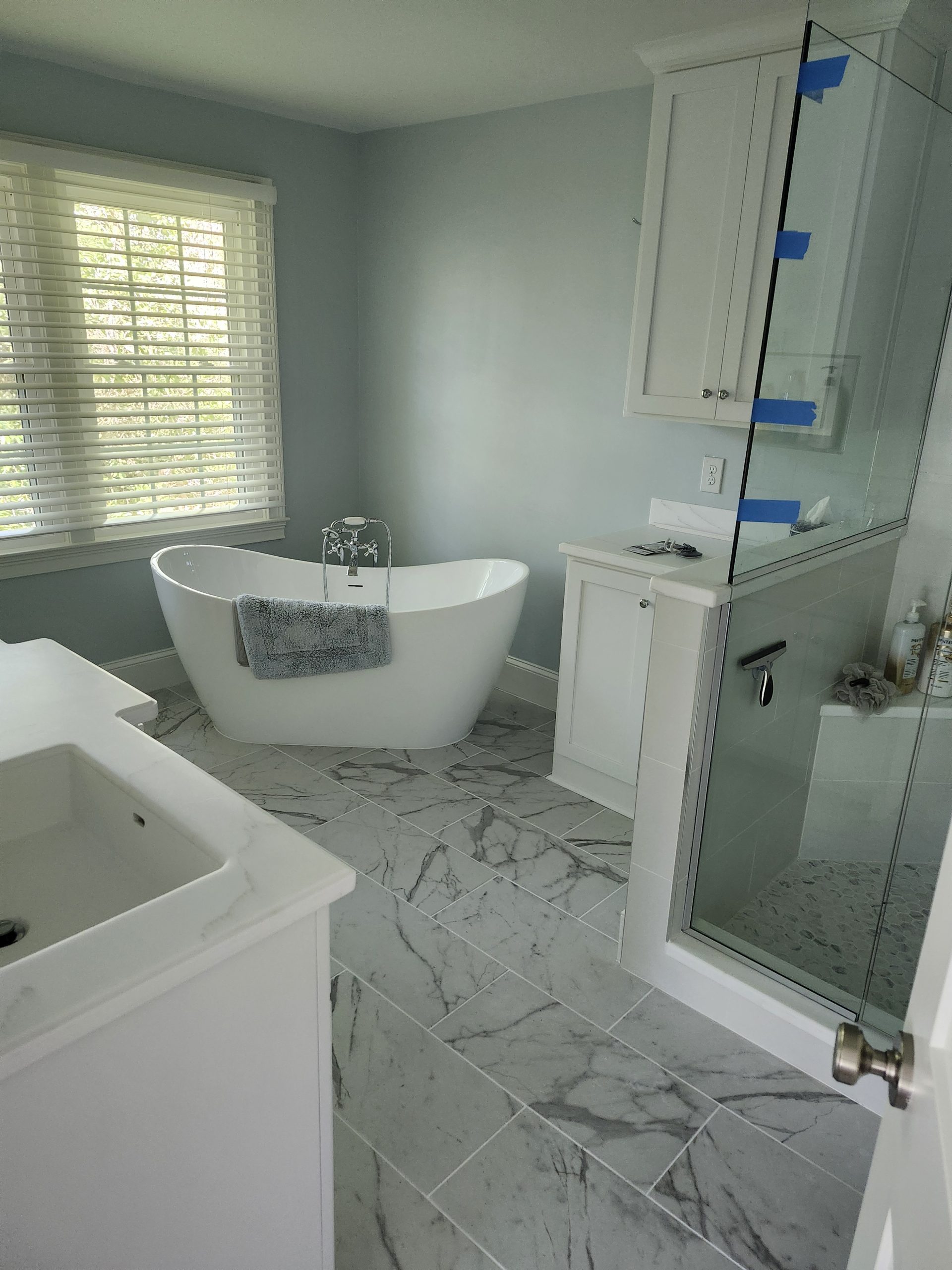 Modern bathroom renovation with freestanding soaking tub, marble tile flooring, frameless glass shower, and custom white cabinetry in progress.