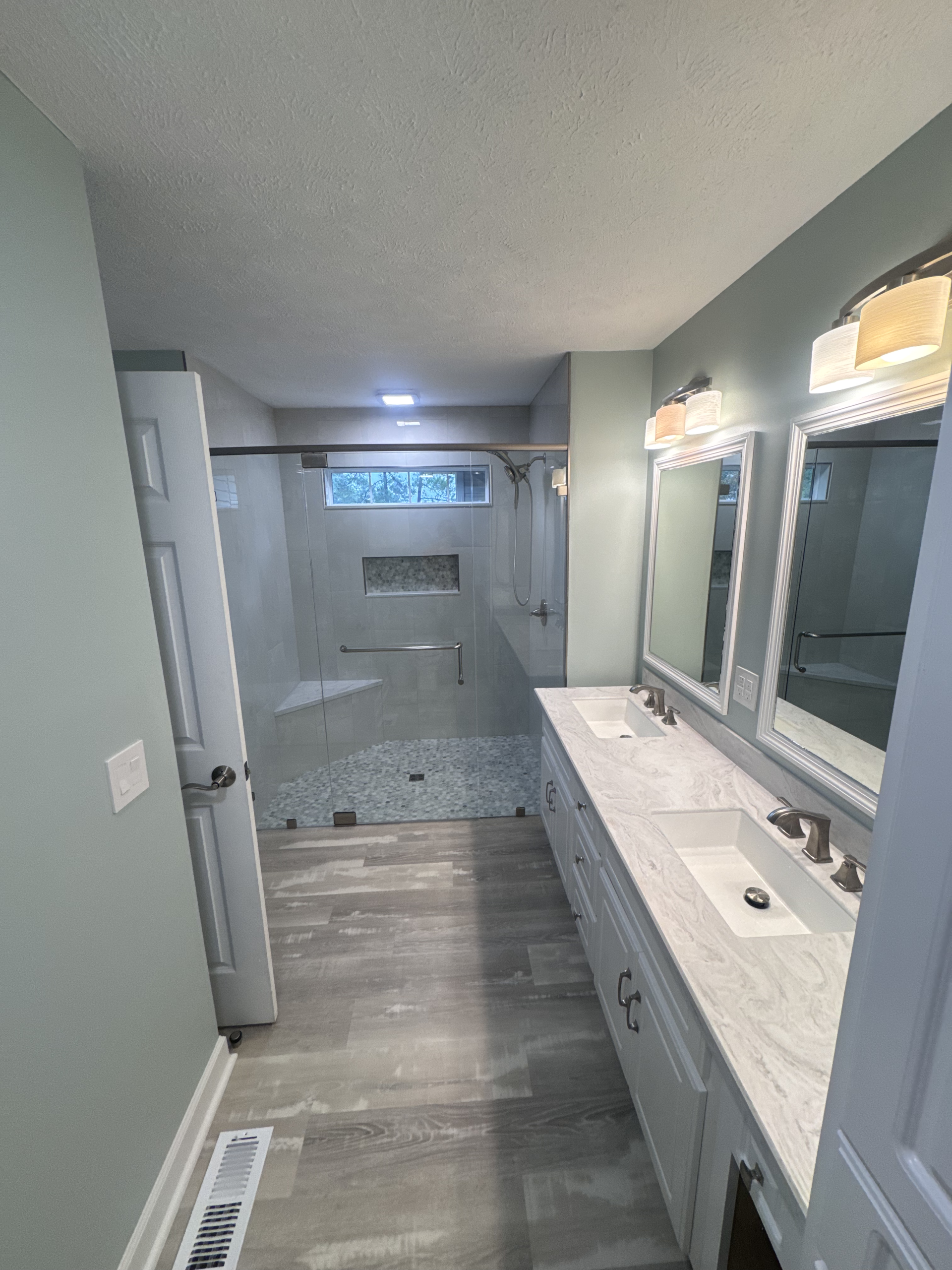Modern bathroom renovation with marble double vanity, glass-enclosed shower, and contemporary fixtures in soft gray-green palette.